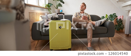 Portrait of young man sitting in living room, dreaming about holiday, sitting with suitcase and smartphone, looking aside thoughtful 126014540