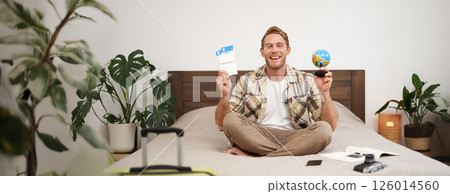 Portrait of cheerful, happy tourist, man sitting with plane tickets and a globe, packed suitcase for travelling around the world, going on vacation, excited about his holiday Portrait of cheerful, happy tourist, man sitting with plane tickets and a globe, packed suitcase for travelling around the world, going on vacation, excited about his holiday 126014560