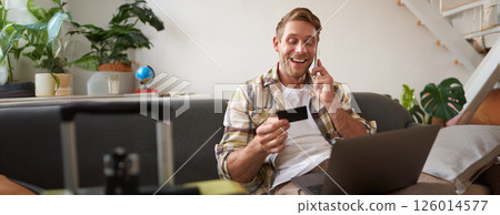 Portrait of young man with credit card, sitting and using laptop, making a phone call, booking hotel or tickets, confirming his purchase over telephone, has suitcase near him 126014577