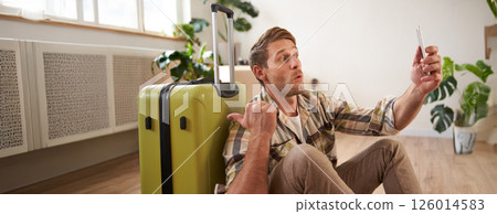 Portrait of happy tourist, man takes selfies before his flight, photos with suitcase, uses smartphone to post pictures on social media 126014583