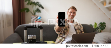 Portrait of handsome young man, showing smartphone screen, recommending mobile app for travelling or booking online, sitting with laptop and suitcase 126014588