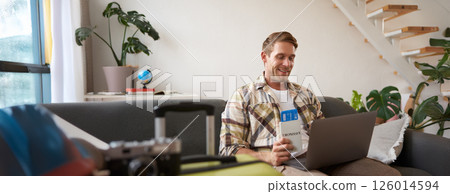 Portrait of handsome young man, has two tickets and passport, goes online on laptop, booking hotel and planning his holiday, sitting in living room Portrait of handsome young man, has two tickets and passport, goes online on laptop, booking hotel and planning his holiday, sitting in living room 126014594