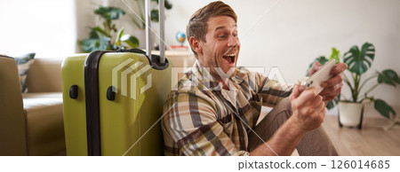 Happy man playing on mobile phone, laughing and smiling, sitting with suitcase, going on holiday, travelling 126014685