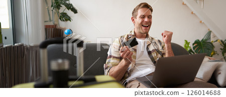 Portrait of happy man, traveller, holding credit card and using laptop, booking vacation, buying tickets online for summer holiday, has suitcase packed Portrait of happy man, traveller, holding credit card and using laptop, booking vacation, buying tickets online for summer holiday, has suitcase packed 126014688