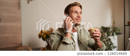 Image of smiling, good-looking young blond man with phone, drinking coffee in cafe, talking to someone over the telephone, answering a call 126014689