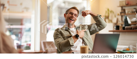 Portrait of handsome, pleased young man shows biceps, flexing muscles, sits in front of laptop in cafe, rejoicing, private coach having a video chat with his client 126014730