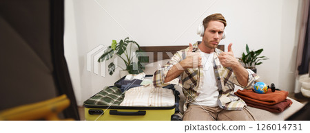 Portrait of handsome man in wireless headphones, shows thumbs up, sits with suitcase, listens to music and packs clothes for vacation, goes on holiday 126014731