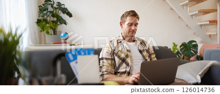 Portrait of handsome young man, has two tickets and passport, goes online on laptop, booking hotel and planning his holiday, sitting in living room Portrait of handsome young man, has two tickets and passport, goes online on laptop, booking hotel and planning his holiday, sitting in living room 126014736