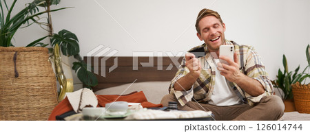 Cheerful tourist, guy with credit card and mobile phone, paying for holiday tickets, sitting on bed with suitcase, packing and going on holiday Cheerful tourist, guy with credit card and mobile phone, paying for holiday tickets, sitting on bed with suitcase, packing and going on holiday 126014744