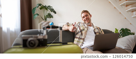 Cheerful man with laptop, tourist sits near suitcase, shows thumb up, books a holiday, goes on vacation Cheerful man with laptop, tourist sits near suitcase, shows thumb up, books a holiday, goes on vacation 126014750