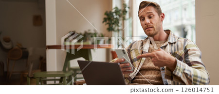 Portrait of young man freelancer, working from cafe, sitting on chair with laptop, pointing finger at his phone screen, showing interest in something on his phone 126014751