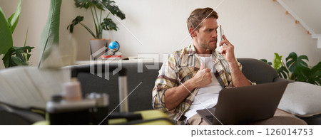 Portrait of angry man waiting on the line, trying to call someone, using smartphone and laptop, sitting near suitcase Portrait of angry man waiting on the line, trying to call someone, using smartphone and laptop, sitting near suitcase 126014753