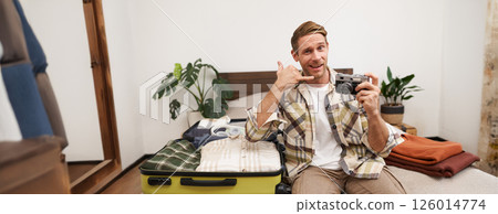Portrait of handsome blond guy with camera in hand, showing phone call gesture and smiling, sitting next to the suitcase, unpacking clothes on vacation, check-in to the hotel on a vacation 126014774