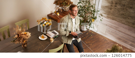 Vertical shot of handsome stylish businessman, standing near the table with laptop, thinking, working on a project on his computer, studying online, doing freelance task 126014775