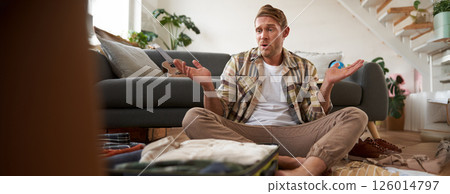 Man packing suitcase, shrugging, deciding what to bring on holiday, looking confused 126014797