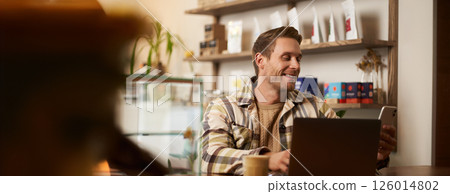 Lifestyle portrait of young businessman, freelancer sitting in cafe, drinking coffee and using laptop, working on online project, digital nomad doing his task, has remote job, looking satisfied 126014802