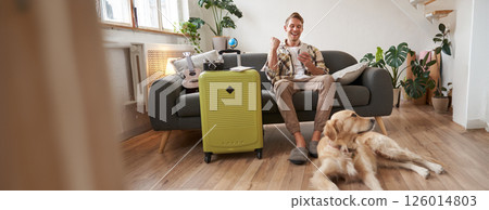 Happy man with his dog, sits in living room with suitcase, looks at smartphone app with excited face, pleased with booked trip 126014803