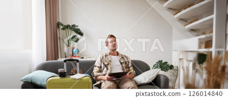 Image of young man, tourist goes on holiday, packed suitcase, sits with digital tablet on sofa, reads, looks at his map, books hotel for business trip 126014840