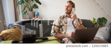 Handsome smiling man with laptop, making a phone call, looking aside while waiting on the telephone, confirms booking online Handsome smiling man with laptop, making a phone call, looking aside while waiting on the telephone, confirms booking online 126014865