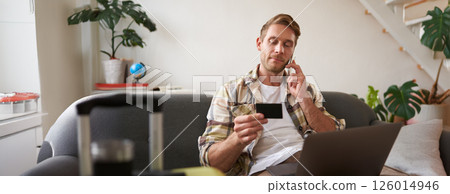 Portrait of young man with credit card, sitting and using laptop, making a phone call, booking hotel or tickets, confirming his purchase over telephone, has suitcase near him Portrait of young man with credit card, sitting and using laptop, making a phone call, booking hotel or tickets, confirming his purchase over telephone, has suitcase near him 126014946