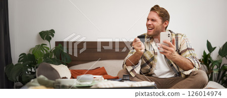 Cheerful male tourist, man packing suitcase, paying for tickets with credit card and smartphone app, celebrating, excited about going on holiday 126014974