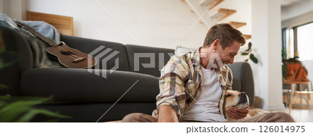 Portrait of happy young man, hugging and playing with his dog, sitting on floor with sofa, looking at golden retriever Portrait of happy young man, hugging and playing with his dog, sitting on floor with sofa, looking at golden retriever 126014975