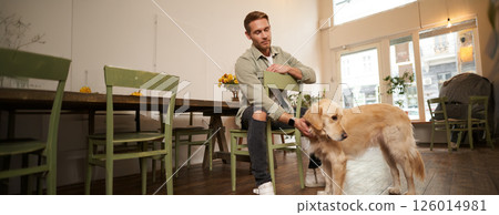 Close up portrait of handsome young man waiting for his order in coffee shop, petting a dog, resting in pet-friendly cafe 126014981