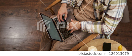 Upper angle cropped shot of male hands, man sitting in cafe with cup of coffee and working on laptop, typing on keyboard, has a phone on the table 126015079