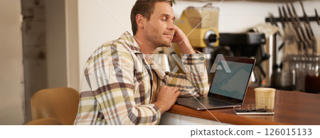 Close up portrait of handsome man, sitting at counter and looking at laptop screen with pleased smile, drinking coffee, browsing on the internet, watching something on computer 126015133