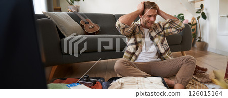 Portrait of man trying to decide what to bring on holiday, packing suitcase, looking in panic at his luggage, going on vacation 126015164