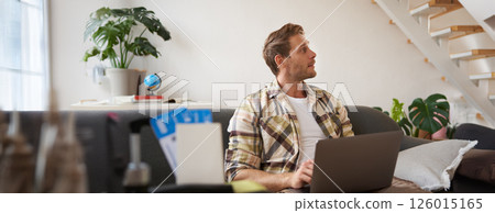 Portrait of young handsome man sitting with laptop, has passport and two plane tickets prepared, planning his journey on computer, booking hotels and restaurants online for the trip 126015165
