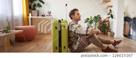 Portrait of handsome young man, tourist sitting with his suitcase, holding passport and flight tickets, going on holiday 126015167