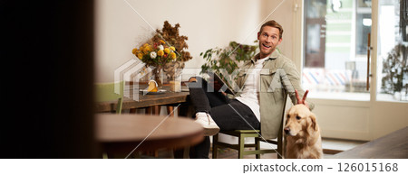 Portrait of happy smiling young man, cafe visitor, sitting in coffee shop with his dog, petting golden retriever, holding digital tablet, reading news or browsing on internet Portrait of happy smiling young man, cafe visitor, sitting in coffee shop with his dog, petting golden retriever, holding digital tablet, reading news or browsing on internet 126015168