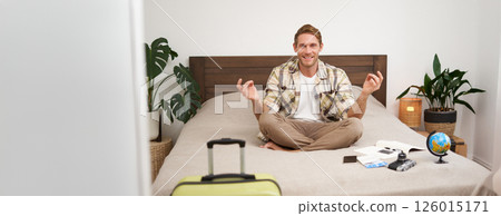 Portrait of young man meditating, packing suitcase, sitting on bed with globe, camera and plane tickets, relaxing before travelling abroad 126015171