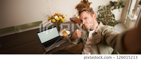 Portrait of handsome man making angry threatening face for selfie photo, taking picture on smartphone app in cafe, sitting near laptop 126015213