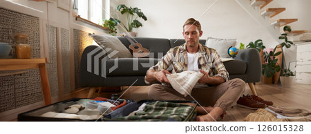 Portrait of young man picking clothes for holiday, sitting on floor with suitcase, packing on vacation, going on a business trip 126015328