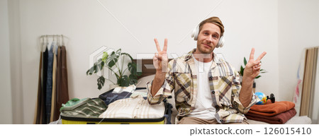 Portrait of cheerful handsome man, tourist going on vacation, dancing and listening to music in headphones while packing suitcase with clothes for holiday trip abroad 126015410