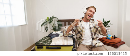 Handsome blond man in headphones, sits on bed with suitcase and clothes, listens to music in wireless headphones while packing for a holiday, going on trip 126015415