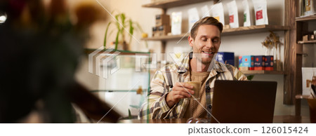 Lifestyle portrait of young businessman, freelancer sitting in cafe, drinking coffee and using laptop, working on online project, digital nomad doing his task, has remote job, looking satisfied 126015424