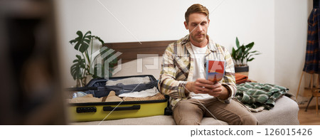 Portrait of young man, tourist is packing his luggage, looking at tour guide, travel map, sitting on bed with his clothes in a suitcase Portrait of young man, tourist is packing his luggage, looking at tour guide, travel map, sitting on bed with his clothes in a suitcase 126015426