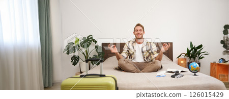 Portrait of young relaxed tourist, man sitting on bed with crossed legs and meditating, has packed luggage, globe and camera, going on holiday, calms down before going on a trip 126015429