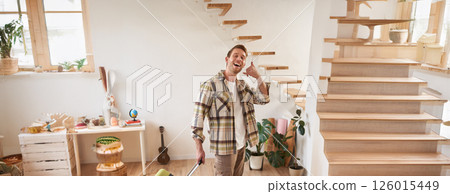 Vertical portrait of happy young man going on holiday, shows mobile phone call hand gesture, walking with suitcase, leaving rented apartment 126015449
