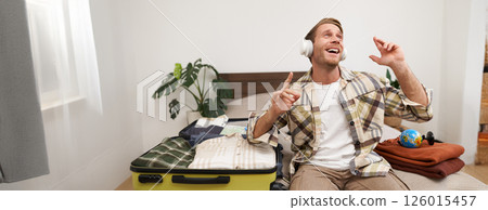 Portrait of cheerful handsome man, tourist going on vacation, dancing and listening to music in headphones while packing suitcase with clothes for holiday trip abroad 126015457