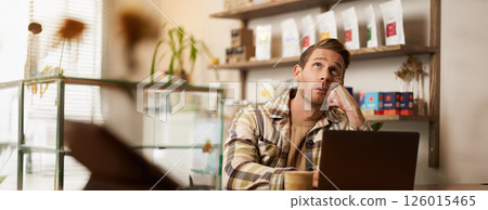 Portrait of stressed working man, sitting in cafe with laptop, business owner doing project online, looking up, roll his eyes, drinking coffee in co-working creative space 126015465