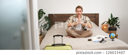 Portrait of young mindful man, tourist going on retreat vacation, packing suitcase, preparing for relaxing vacation, meditating, sitting on bed with globe, plane tickets and camera 126015487