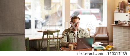 Image of young digital nomad, man in glasses sits in cafe, works from coffee shop, uses laptop in co-working space, wears glasses, drinks his beverage 126015489