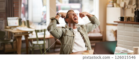 Portrait of pleased, smiling young man shouting with rejoice, sitting in cafe in front of laptop, flexing muscles, screaming from happiness, winning and celebrating Portrait of pleased, smiling young man shouting with rejoice, sitting in cafe in front of laptop, flexing muscles, screaming from happiness, winning and celebrating 126015546