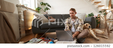 Portrait of man with dog, sitting on floor, has suitcase and laptop opened, booking or buying online, planning holiday, packing for vacation Portrait of man with dog, sitting on floor, has suitcase and laptop opened, booking or buying online, planning holiday, packing for vacation 126015592