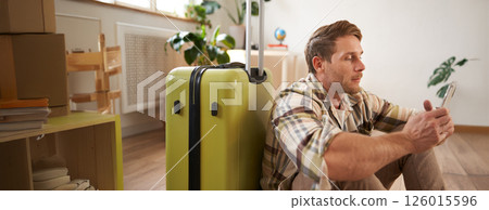 Portrait of young man, traveller sits with suitcase and mobile phone, chats on smartphone 126015596