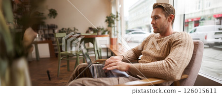 Working people and public space concept. Young handsome man, ux ui designer sitting with laptop in cafe, drinking cappuccino, looking at screen with serious face Working people and public space concept. Young handsome man, ux ui designer sitting with laptop in cafe, drinking cappuccino, looking at screen with serious face 126015611